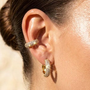 May include: A gold ear cuff and a matching gold hoop earring, both featuring a thick, rounded band encrusted with numerous small, sparkling clear stones that catch the light, creating a bright, reflective, and textured metallic appearance.