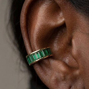 May include: A gold ear cuff featuring rectangular, emerald-green gemstones. The cuff is worn on the ear, highlighting the vivid green stones. The jewellery piece adds a touch of elegance and colour.