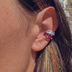 May include: Ear cuff jewelry featuring two stacked bands. The top band is white with rectangular stones, and the bottom band is pink with rectangular stones. The jewelry is worn on the ear.
