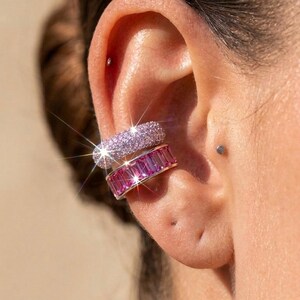 May include: Two stacked ear cuffs worn on the cartilage. The top cuff is covered in small, light pink pavé stones. The bottom cuff features a row of rectangular, deep pink baguette-cut stones set in a polished silver-toned metal band.