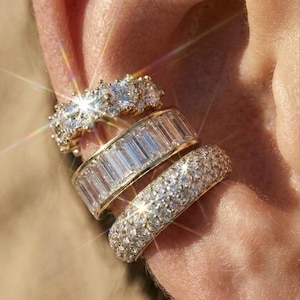 Pode incluir: Close-up de três brincos de ouro adornados com pedras preciosas brilhantes. O brinco superior apresenta pedras redondas, o do meio tem pedras retangulares e o inferior é coberto por pequenas pedras preciosas brilhantes.