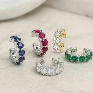 May include: Five ear cuff earrings with various gemstones. The earrings include blue sapphires, rubies, diamonds, emeralds, and a gold cuff with diamonds. The gemstones are set in a row along the curved design.