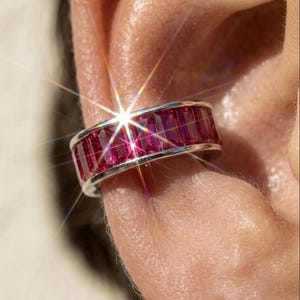 May include: A silver-toned ear cuff featuring a row of rectangular, baguette-cut deep pink stones set within a polished metal channel. The jewellery piece is positioned on the outer edge of an ear, reflecting light with a prominent starburst effect.