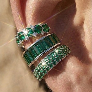 Può includere: Tre ear cuff color argento con pietre verdi. Il modello superiore presenta pietre verdi rotonde con piccoli accenti chiari, quello centrale ha pietre verdi rettangolari taglio smeraldo e quello inferiore è tempestato di piccole pietre verdi rotonde.