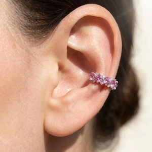 May include: A silver-toned ear cuff featuring a row of four round, vibrant pink gemstones alternating with smaller, clear white stones. The jewelry piece is positioned securely on the cartilage of the ear, showcasing a polished metal band.