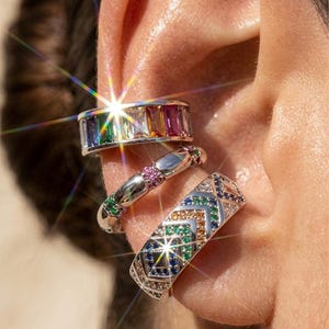 Peut inclure: Trois manchettes d'oreille argentées ornées de pierres précieuses colorées. La manchette supérieure présente un arc-en-ciel de pierres rectangulaires. La manchette du milieu a des pierres roses et violettes. La manchette inférieure a un motif géométrique.