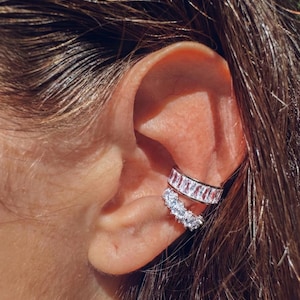 Peut inclure: Gros plan sur un ear cuff argenté avec deux rangées de pierres de zircone cubique claires et étincelantes. La rangée supérieure présente des pierres rectangulaires, tandis que la rangée inférieure comporte des pierres rondes. Le ear cuff est porté sur l'oreille.