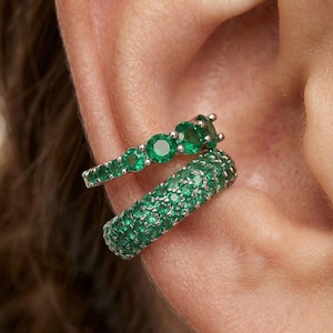 May include: Two ear cuffs with emerald green gemstones. One cuff features a row of round emeralds, while the other is covered in smaller emeralds. The jewelry is set against the backdrop of a person's ear.