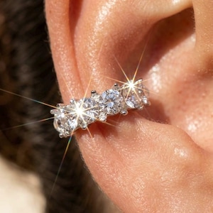 May include: A silver-toned ear cuff featuring a row of five round, clear, faceted stones set in a prong setting. The stones exhibit a high-shine, reflective quality with visible light flares, creating a bright, sparkling appearance on the ear cartilage.