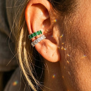 Peut inclure: Deux bagues d'oreille portées sur le cartilage. La bague supérieure présente une rangée de cinq pierres rondes vertes serties sur une bande métallique. La bague inférieure est une bande incrustée d'une rangée de pierres claires et brillantes.
