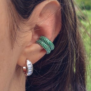 Op de afbeelding: Close-up van een oor versierd met twee oormanchetten en een oorring. De oormanchetten zijn bezet met kleine, levendige groene stenen. De oorring heeft rechthoekige, heldere stenen gezet in zilver metaal.