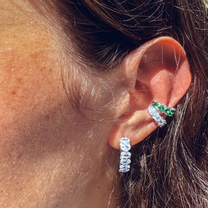Può includere: Primo piano di un orecchio adornato da due gioielli. Un piccolo orecchino a cerchio con pietre trasparenti di forma ovale è nel lobo. Un orecchino a polsino con pietre bianche e verdi è sulla parte superiore dell'orecchio.