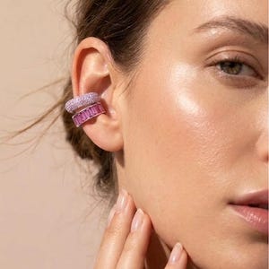 Könnte beinhalten: Zwei Ohrklemmen am oberen Knorpelbereich des Ohrs. Die obere Klemme ist ein breiter Ring, der mit kleinen, hellvioletten Pavé-Steinen besetzt ist. Die untere Klemme ist ein breiterer Ring mit einer Reihe rechteckiger, rosafarbener Baguette-Steine.