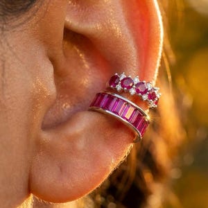 Könnte beinhalten: Nahaufnahme eines Ohrs, das mit zwei Ohrklemmen geschmückt ist. Die untere Manschette hat rechteckige, leuchtend rote Steine, während die obere Manschette eine Reihe von runden roten Steinen mit kleineren weißen Steinen aufweist. Schmuck.