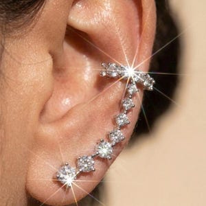 May include: A silver-toned ear crawler earring featuring a curved row of seven round, clear, faceted stones that follow the natural shape of the ear cartilage. The metal setting is polished and reflective, creating a bright, sparkling appearance.