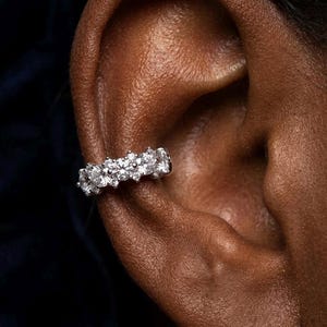 May include: A silver-toned ear cuff featuring a row of five round, clear, faceted stones set in a metallic band. The jewellery piece is positioned securely along the cartilage of the ear, showcasing a bright, reflective finish and a cluster design.