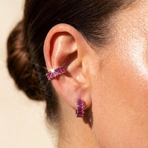 May include: A close-up of an ear adorned with a pink gemstone ear cuff and matching hoop earrings. The jewelry features rectangular-cut pink stones set in a silver-toned metal. The ear cuff wraps around the upper ear, while the earrings are a small hoop design.