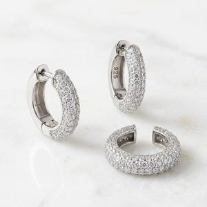 Può includere: Due orecchini a cerchio in argento e un ear cuff, ciascuno caratterizzato da un'incastonatura pavé di piccole pietre trasparenti. La superficie metallica degli orecchini a cerchio reca il numero 925, che indica la composizione in argento.