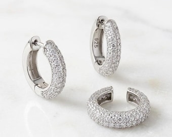 Pode incluir: Dois brincos de argola em prata e um ear cuff, cada um com uma cravação pavé de pequenas pedras transparentes. A superfície metálica das argolas apresenta o número 925 gravado, indicando a composição em prata de lei destas peças.