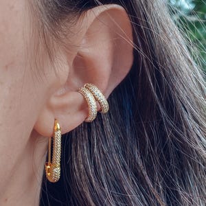 Op de afbeelding: Goudkleurige veiligheidsspeld oorbellen en oor manchet set. De oorbellen en oor manchet zijn bezet met kleine, sprankelende steentjes. De veiligheidsspeld oorbel hangt aan de oorlel, terwijl de oor manchet om het bovenste oor wikkelt.