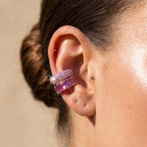 Puede incluir: Dos pendientes tipo ear cuff en el cartílago. El superior es una banda gruesa cubierta de pequeñas piedras rosas brillantes. El inferior es una banda más ancha con una fila de piedras rosas rectangulares de corte baguette en un marco metálico.
