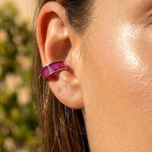 Può includere: Primo piano di un orecchino a cerchio d'argento ornato di pietre preziose rosa rettangolari. Il gioiello è indossato sull'orecchio, mostrando il design e il colore sulla pelle. Lo sfondo è sfocato, evidenziando l'orecchino.