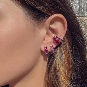 Puede incluir: Primer plano de una oreja adornada con tres joyas. Las joyas incluyen un par de pendientes de aro pequeños, un brazalete de oreja de doble banda y un par de pendientes de botón, todos con piedras rectangulares de color rubí.