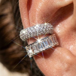 May include: Two silver-toned ear cuffs worn on the cartilage. The top cuff features a dense pavé setting of small, sparkling clear stones. The bottom cuff is designed with a row of rectangular, baguette-cut clear stones set in a smooth metal band.