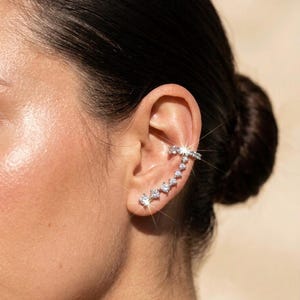 May include: A silver-toned ear cuff featuring a curved row of sparkling, clear round-cut stones that follow the contour of the ear. The jewelry piece consists of a horizontal bar at the top connected to a descending line of individual prong-set crystals.