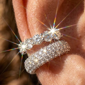 May include: Two silver-toned ear cuffs worn on the ear. The top cuff features a row of five large, round, clear, faceted stones. The bottom cuff is a wider band densely encrusted with numerous small, round, clear, faceted stones that sparkle.