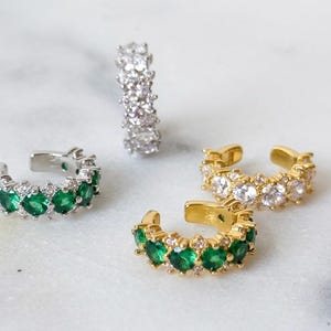May include: Four ear cuffs arranged in a cluster. One silver-toned cuff features clear stones, another silver-toned cuff features green stones, and two gold-toned cuffs feature either clear or green stones. Each cuff has a circular open band design.