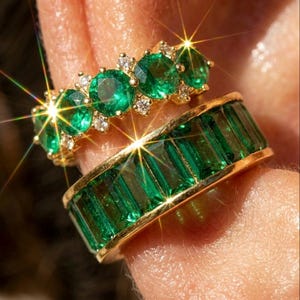 Könnte beinhalten: Zwei goldene Ringe mit Smaragden. Der obere Ring hat runde Smaragde und kleine Diamanten. Der untere Ring hat rechteckige Smaragde. Beide Ringe haben ein goldenes Band. Die Ringe befinden sich an einem Finger.