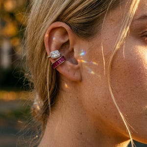 Pode incluir: Dois brincos tipo ear cuff usados na cartilagem. O superior apresenta uma pedra retangular transparente rodeada por um halo de pequenas pedras brancas. O inferior é uma banda cravejada com uma fila de pedras retangulares rosa-escuro.