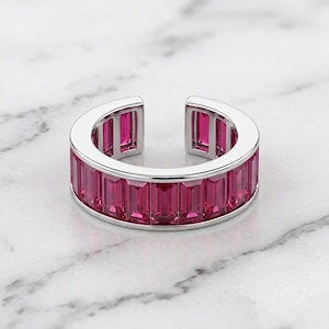 Könnte beinhalten: Ein silberfarbener offener Ring mit einer durchgehenden Reihe rechteckiger, im Baguetteschliff geschliffener, leuchtend pinker Steine entlang des äußeren Umfangs. Der Ring hat eine polierte Metalloberfläche und eine Öffnung an der Oberseite.
