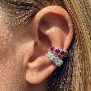 May include: Two ear cuffs are displayed on an ear. The upper cuff has five red gemstones, while the lower cuff is adorned with white gemstones. The ear is set against a backdrop of blonde hair.