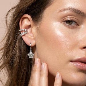 May include: Silver ear cuff and star-shaped hoop earrings with clear gemstones. The ear cuff wraps around the upper ear, while the earrings dangle below. The jewelry is set against a soft, neutral background.