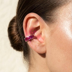 Può includere: Un ear cuff color argento caratterizzato da una fila di pietre preziose rettangolari color rosa intenso. La fascia metallica trattiene le pietre in un'incastonatura a binario, creando una linea di colore continua che avvolge la cartilagine.