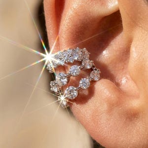 Peut inclure: Une boucle d'oreille de type ear cuff argentée composée de trois anneaux superposés ornés de multiples pierres rondes, transparentes et facettées. Le bijou présente une finition métallique brillante et un éclat lumineux intense.