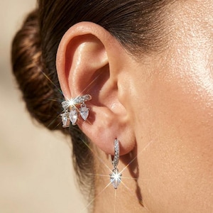 Peut inclure: Gros plan sur une manchette d'oreille et une boucle d'oreille en argent. La manchette d'oreille présente une rangée de petites pierres transparentes avec trois pierres en forme de poire pendantes. Bijoux.