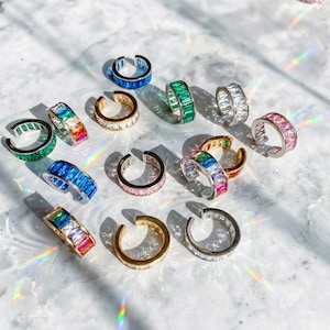 以下が含まれることがあります： 青、緑、ピンク、虹色など、さまざまな色のイヤーカフのコレクション。長方形にカットされた宝石が飾られており、白い表面に展示されています。