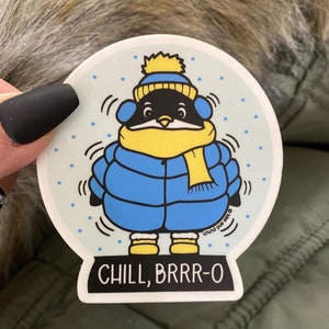 Könnte beinhalten: Runder Aufkleber mit einem Cartoon-Pinguin in einer blauen Steppjacke, einem gelben Schal und einer Mütze mit Ohrenschützern. Der Text "CHILL, BRRR-O" steht unten. Der Pinguin ist von blauen Schneeflocken umgeben.
