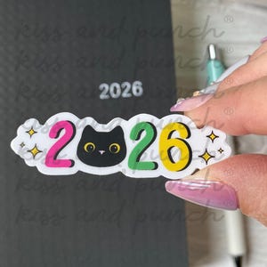 May include: A clear sticker with the year "2026" in colorful text and a black cat face. The numbers are pink, green, and yellow, with star accents. The sticker is held in front of a dark gray notebook.