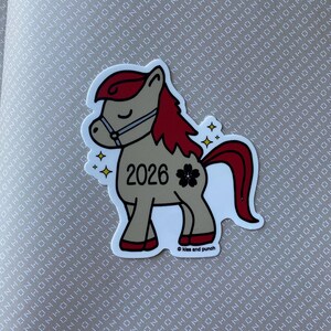May include: A sticker depicting a cartoon pony with a tan body, red mane and tail, and a bridle. The pony has the number "2026" and a black flower on its side. The sticker has a white border and the text "Kiss and punch".