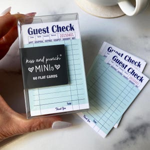Puede incluir: Una caja de plástico transparente que contiene blocs de cheques para invitados. Los blocs tienen un patrón de cuadrícula azul claro y las palabras "Guest Check" impresas en azul oscuro. También se incluye una tarjeta negra con el texto "kiss and punch MINIs".