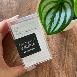 Könnte beinhalten: Eine durchsichtige Kunststoffbox mit 60 flachen Karten im Design von alten Bibliotheksausleihkarten. Die schwarze Papierbanderole trägt den weißen Text kiss and punch MINIs 60 FLAT CARDS mit Herzsymbolen. Die Karten haben Zeilen für Autor, Titel, Name des Entleihers und Fälligkeitsdatum.