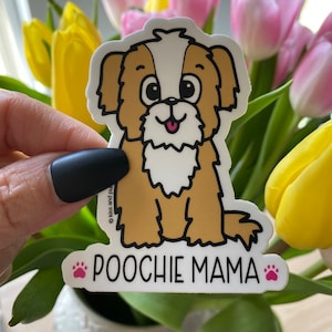 May include: A sticker featuring a cartoon dog with brown and white fur, a pink tongue, and large eyes. The sticker reads "POOCHIE MAMA" in black text with a pink paw print. The sticker is held in front of a bouquet of flowers.