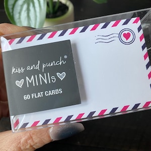 Könnte beinhalten: Eine transparente Kunststoffhülle mit flachen Karten. Die Karten haben einen weißen Hintergrund mit einem rosa und marineblauen Rand. Eine kleine schwarze Karte mit dem Text "kiss and punch MINIs" und "60 FLAT CARDS" ist sichtbar.