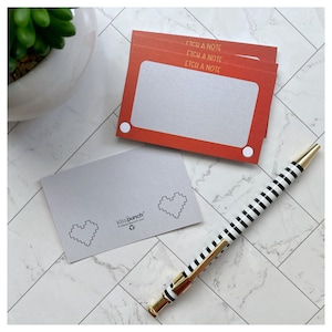 Pode incluir: Uma pilha de blocos de notas vermelhos com o texto "Etch a Note" e um design retangular branco, que lembra um brinquedo retrô. Um bloco de notas cinza com gráficos de coração pixelados e uma caneta listrada em preto e branco com detalhes dourados.
