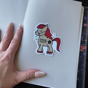May include: A sticker of a cartoon horse with a red mane and tail, wearing a bridle. The sticker has the year "2026" and a flower graphic. The sticker is on a notebook with a patterned cover.