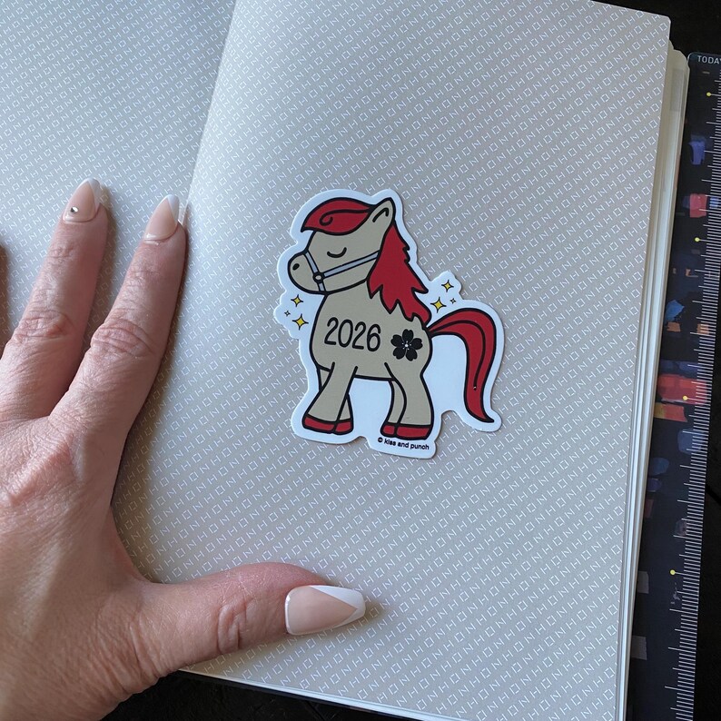 May include: A sticker of a cartoon horse with a red mane and tail, wearing a bridle. The sticker has the year "2026" and a flower graphic. The sticker is on a notebook with a patterned cover.